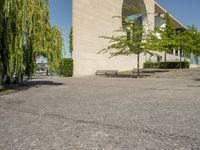 a paved walkway next to a building and a large tree in front of it, with a stone slab sitting in the middle