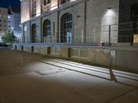 the sidewalk has shadows across it on the side of the street at night with buildings lit up in the background