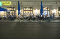 there are a lot of people sitting outside of a restaurant at night eating food on the sidewalk