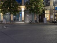a city street lined with stores and trees at night with a bike rack nearby from a cafe