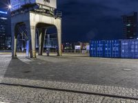 a train station lit up at night, with blue containers in the background, and a yellow tower near by