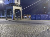 a train station lit up at night, with blue containers in the background, and a yellow tower near by