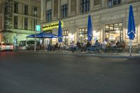 a restaurant with patio umbrellas next to the street at night time as people sit outside