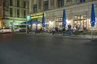 a restaurant with patio umbrellas next to the street at night time as people sit outside