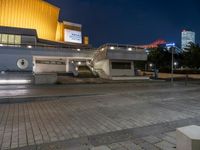 a building with a stairway next to it at night, in an empty plaza with lights shining off and buildings in the background