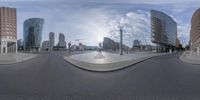 a panoramic view of a city street with tall buildings and a fountain in the foreground