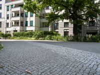 the road is cobble stone between two buildings and a sidewalk in front of a tree and bushes