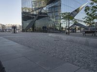 large mirrored building reflecting trees in a city park plaza with a few benches at the end