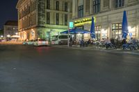 cars parked outside an outdoor restaurant at night with people dining outside the building and tables lined up in front of it