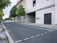 a paved street with a grey building on the side, next to two trees and grass