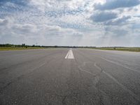 an airport runway with empty roadway and clouds overhead on blue and grey sky day, with plane taking off from the airport