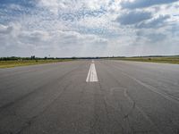 an airport runway with empty roadway and clouds overhead on blue and grey sky day, with plane taking off from the airport