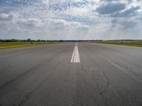 an airport runway with empty roadway and clouds overhead on blue and grey sky day, with plane taking off from the airport