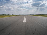 an airport runway with empty roadway and clouds overhead on blue and grey sky day, with plane taking off from the airport