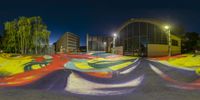 a skateboard park with skate board ramps and buildings at night time, all illuminated by street lights