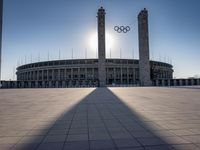 a stadium and the olympic symbol and a flag are on display outside it in front of the sun