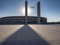 a stadium and the olympic symbol and a flag are on display outside it in front of the sun