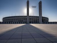 a stadium and the olympic symbol and a flag are on display outside it in front of the sun