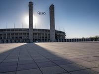 a stadium and the olympic symbol and a flag are on display outside it in front of the sun