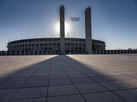 a stadium and the olympic symbol and a flag are on display outside it in front of the sun