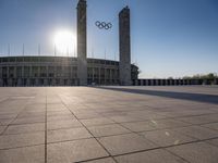 a stadium and the olympic symbol and a flag are on display outside it in front of the sun