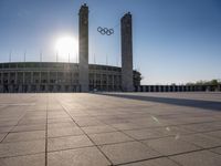 a stadium and the olympic symbol and a flag are on display outside it in front of the sun