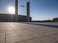 a stadium and the olympic symbol and a flag are on display outside it in front of the sun