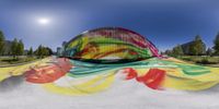 a large round object painted on to a wall in a park area with a big rainbow colored sculpture in the foreground