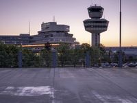 two cars parked in a parking lot beside a control tower and other buildings at dusk