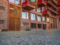 the brick walkway next to the red building is lined with windows and potted plants