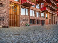 the brick walkway next to the red building is lined with windows and potted plants