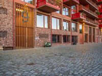 the brick walkway next to the red building is lined with windows and potted plants