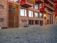 the brick walkway next to the red building is lined with windows and potted plants