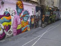 several people walking past colorful wall paintings in a narrow alley path between two buildings with parked cars