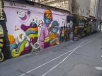 several people walking past colorful wall paintings in a narrow alley path between two buildings with parked cars