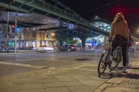 a woman with a bike stands on a city street during the night with an urban bridge above