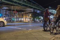 a woman with a bike stands on a city street during the night with an urban bridge above