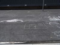 a group of chalk drawings on pavement near a metal fence area where a person is walking with another person