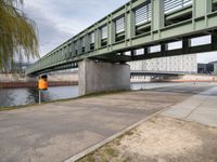 the yellow bin under the bridge is near the water's edge and the sidewalk and the path