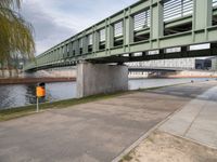 the yellow bin under the bridge is near the water's edge and the sidewalk and the path