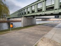 the yellow bin under the bridge is near the water's edge and the sidewalk and the path