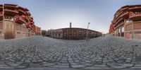 a 360 - shot lens shows the view of a building from one side of a cobblestone street in front of a red brick building