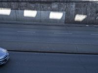 cars and a truck drive by a concrete wall on a street with multiple orange cones