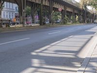 a side walk that shows two cars parked on the street, and below a bridge