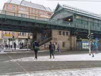 people are walking across the snow in front of a green bridge and several buildings and signs