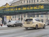 cars drive on the street at a bus stop in europe with an overpass and green bridge over it