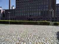 a brick courtyard is pictured with tall buildings in the background from which trees and bushes are planted near the benches