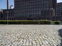 a brick courtyard is pictured with tall buildings in the background from which trees and bushes are planted near the benches