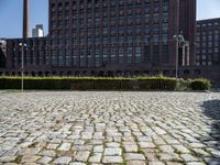 a brick courtyard is pictured with tall buildings in the background from which trees and bushes are planted near the benches