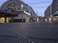 an empty, well - lit city street at dusk with people walking by the buildings